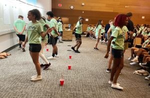 10-15 students, all dressed in green Honors Program shirts, playing a game linking them by string to red cups.