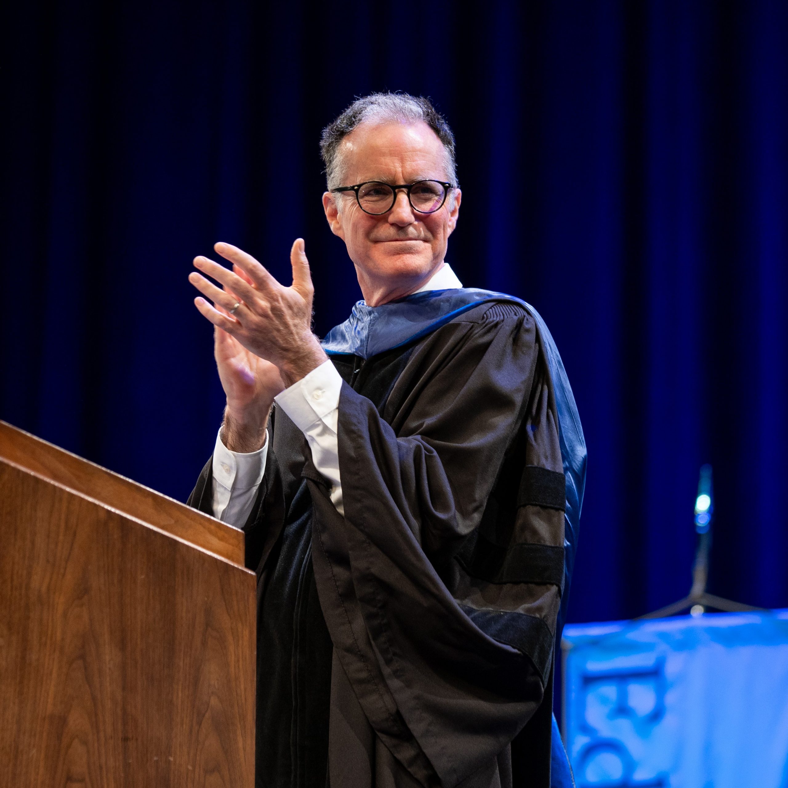 Ken Cormier on stage at the 2025 Honors Medals Ceremony, clapping while wearing black academic regalia