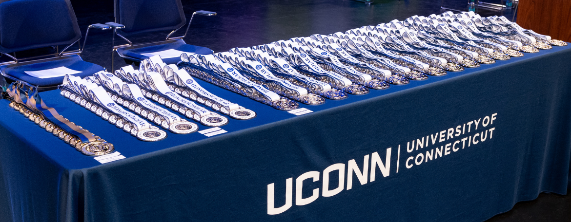Dozens of silver and gold accented medals attached to white ribbons on a navy blue tablecloth with a white logo that reads UConn University of Connecticut