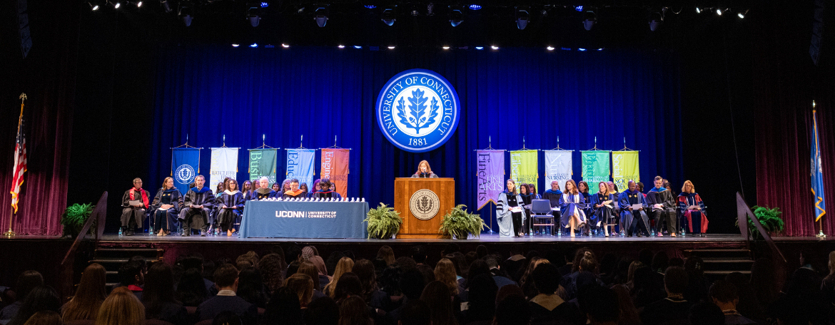 A few dozen UConn faculty and administrators on stage in front of signage for the UConn schools and colleges on either side of a lectern; the lectern has a speaker talking and is below a circular navy blue and white University of Connecticut seal