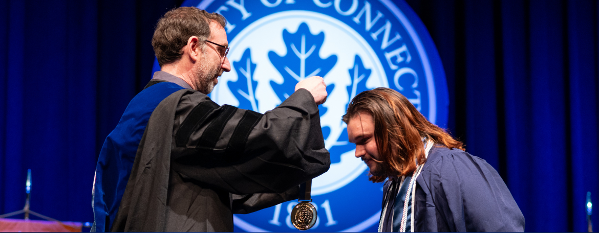 A student in navy blue graduation gown receiving their medal on stage from a faculty member in black academic regalia
