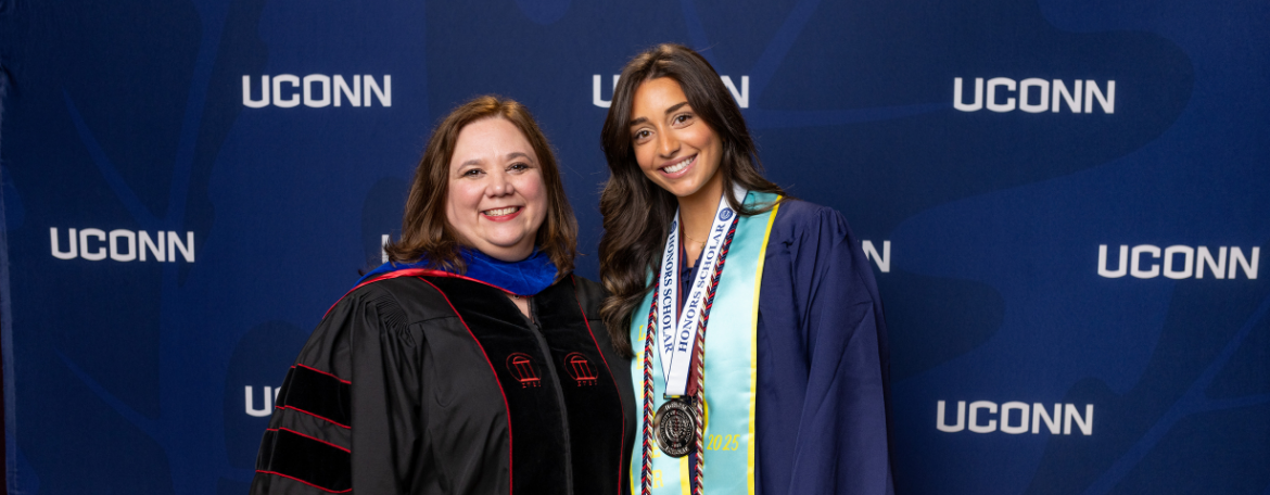 Director of the Honors Program Jennifer Lease Butts is dressed in black academic regalia and posing with a student in a navy blue graduation gown and wearing a medal in front of a navy blue background with white UConn text on it