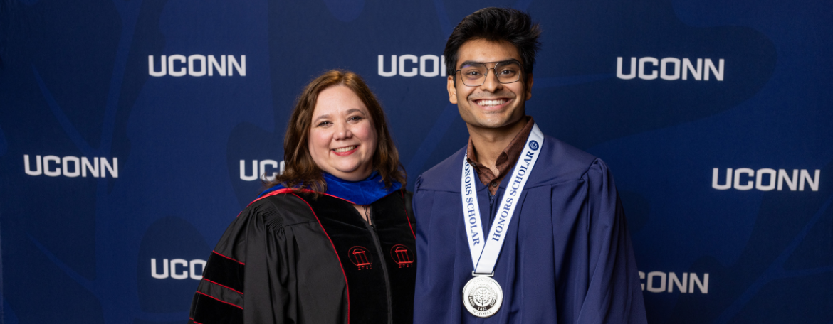 Director of the Honors Program Jennifer Lease Butts is dressed in black academic regalia and posing with a student in a navy blue graduation gown and wearing a medal in front of a navy blue background with white UConn text on it