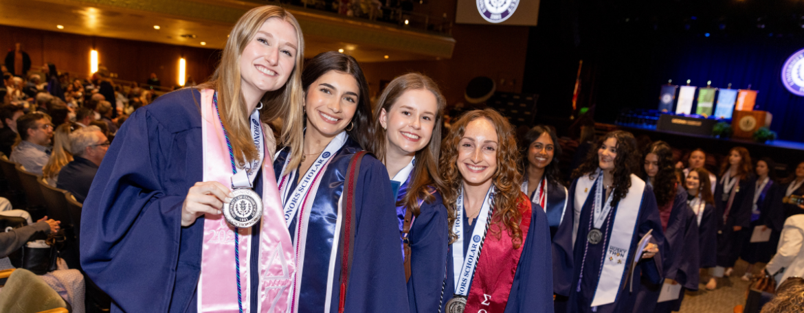 Students in navy blue graduation gowns smiling as they walk down the aisle to leave the Honors Medals Ceremony; 1 is showing off their medal