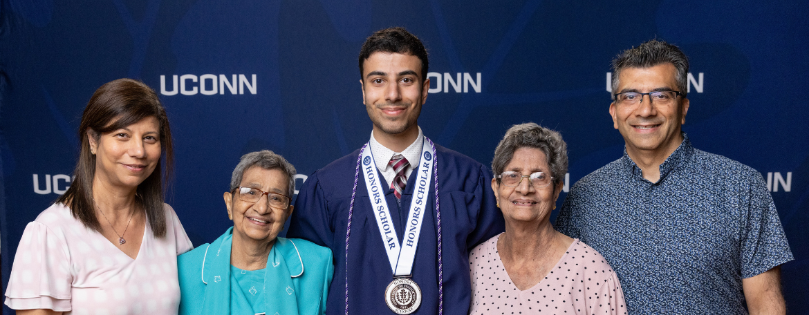 A student in a navy blue graduation gown and wearing a medal in front of a navy blue background with white UConn text on it; the student is with 4 guests