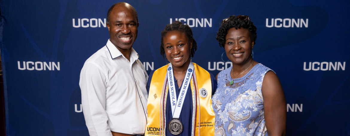 A student in a navy blue graduation gown and wearing a medal in front of a navy blue background with white UConn text on it; the student is with 2 guests