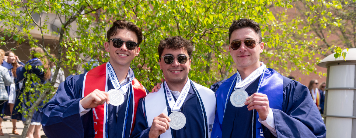 3 students outside in navy blue graduation gowns, each wearing a medal and holding it with 1 hand
