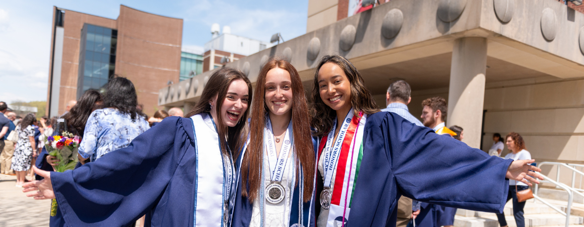 3 students outside in navy blue graduation gowns, each wearing a medal; they are next to each other, and the outer 2 students have their outer arms and hands extended in celebration