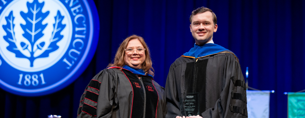 Ryan Talbert receiving the UConn Honors Faculty Member of the Year Award from the Director of the Honors Program Jennifer Lease Butts while both are dressed in black academic regalia