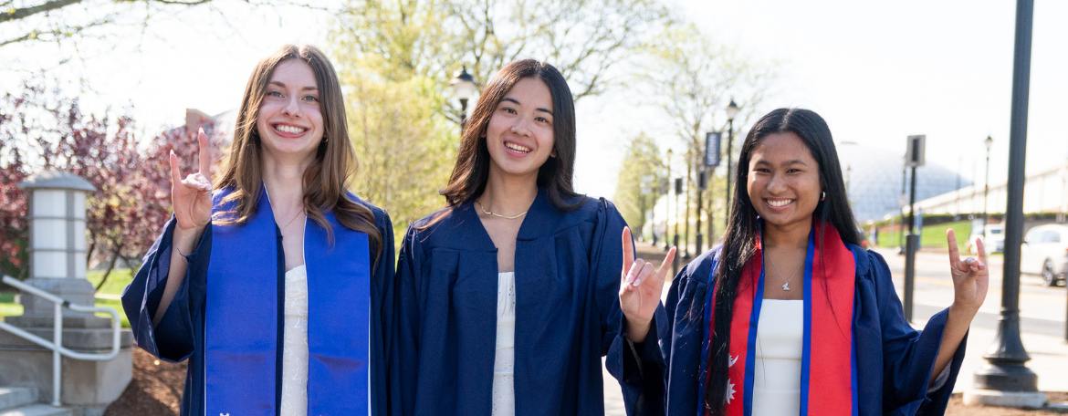 3 students smiling in navy blue graduation gowns outside, and each one is making a UConn Husky dog symbol with 1 of their hands
