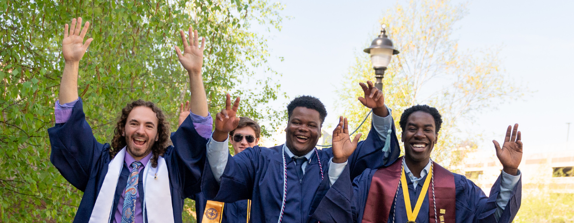4 students in navy blue graduation gowns outside raising their hands in celebration and smiling
