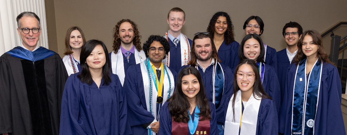 13 University Scholars in navy blue graduation gowns and 1 staff member in black academic regalia smiling in a group