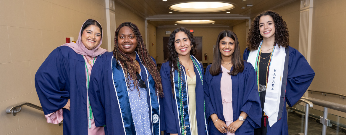 5 students in navy blue graduation gowns smiling with a hallway in the background