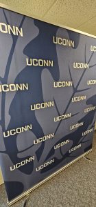 A large navy blue photo backdrop with a faint white oak leaf that repeatedly has UCONN in white letters
