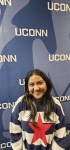 A smiling college student in front of a navy blue photo backdrop with a faint white oak leaf that repeatedly has UCONN in white letters; the student has long black hair and is wait a navy blue and white striped sweater with a large red star in its middle